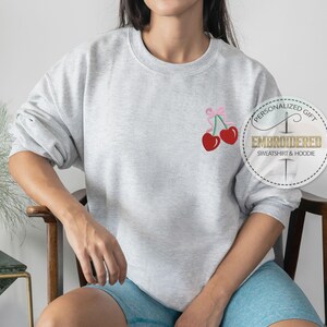 May include: A gray sweatshirt with a pink ribbon and two red cherries embroidered on the front. The sweatshirt has the text "Personalized Gift" and "Embroidered Sweatshirt & Hoodie" on a white circle.