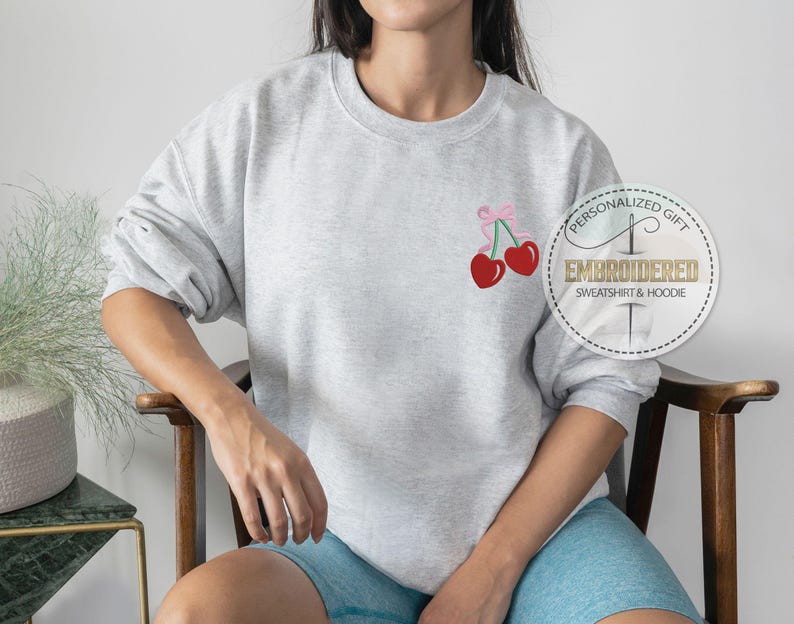 May include: A gray sweatshirt with a pink ribbon and two red cherries embroidered on the front. The sweatshirt has the text "Personalized Gift" and "Embroidered Sweatshirt & Hoodie" on a white circle.