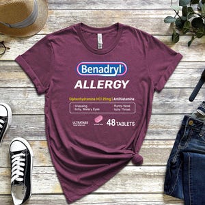 May include: A maroon t-shirt with a white graphic of a Benadryl allergy medication box. The box says "Benadryl Allergy" and "Diphenhydramine HCI 25mg | Antihistamine". The box also lists symptoms like "Sneezing, Itchy, Watery Eyes, Runny Nose, Itchy Throat" and says "48 Tablets".