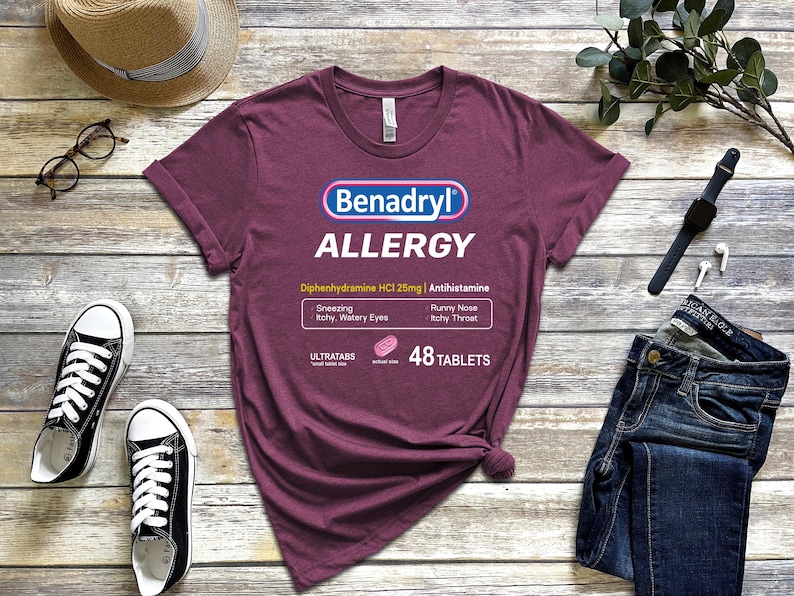 May include: A maroon t-shirt with a white graphic of a Benadryl allergy medication box. The box says "Benadryl Allergy" and "Diphenhydramine HCI 25mg | Antihistamine". The box also lists symptoms like "Sneezing, Itchy, Watery Eyes, Runny Nose, Itchy Throat" and says "48 Tablets".