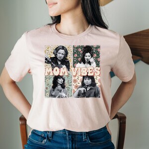 Pu&ograve; includere: Maglietta rosa con una stampa grafica di quattro donne su sfondi floreali. Il testo "MOM VIBES" &egrave; stampato in un font retr&ograve;.