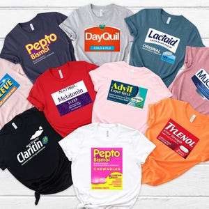 May include: Ten different t-shirts with various medication brand names printed on them. The shirts are in different colors, including pink, red, blue, black, white, and orange. The medication names include Aleve, Pepto Bismol, Claritin, Natrol Melatonin, DayQuil, Lactaid, Advil, Tylenol, Mucinex, and Pepto Bismol Chewables.
