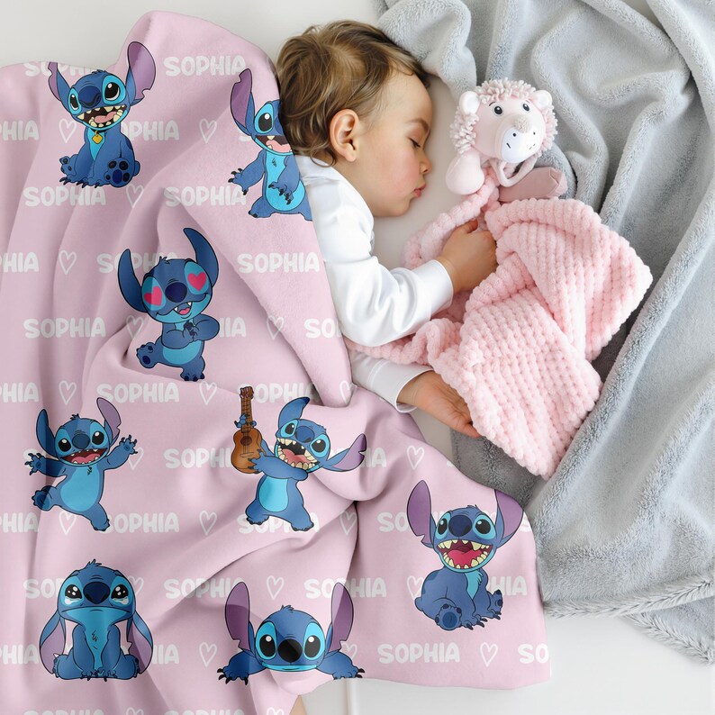 May include: A light pink baby blanket featuring multiple images of a blue cartoon character, hearts, and the name "SOPHIA" in white. A sleeping child is nestled under the blanket, holding a pink plush toy and a gray blanket.