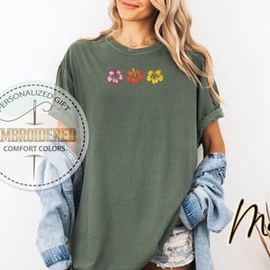 May include: A moss green t-shirt with embroidered hibiscus flowers in pink, orange, and yellow. The shirt has rolled-up sleeves and the text "Moss" in a script font. The shirt also has the text "Personalized Gift" and "Embroidered Comfort Colors".