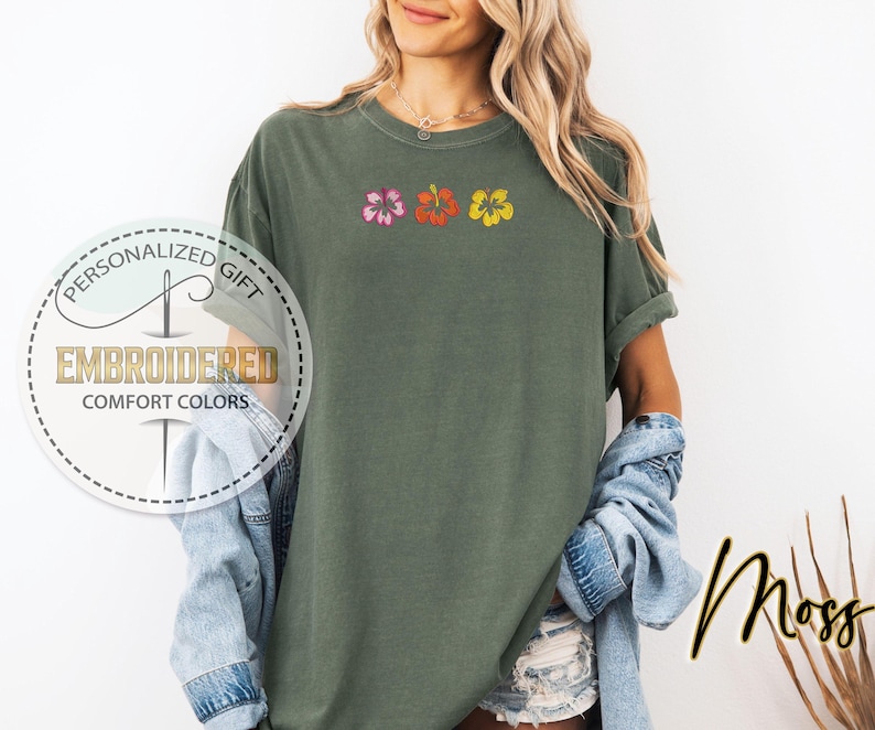 May include: A moss green t-shirt with embroidered hibiscus flowers in pink, orange, and yellow. The shirt has rolled-up sleeves and the text "Moss" in a script font. The shirt also has the text "Personalized Gift" and "Embroidered Comfort Colors".