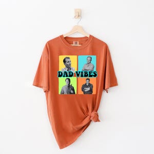 May include: Burnt orange t-shirt with a graphic that says "DAD VIBES". The graphic features four black and white images of men in a grid pattern. The shirt is hanging on a wooden hanger.