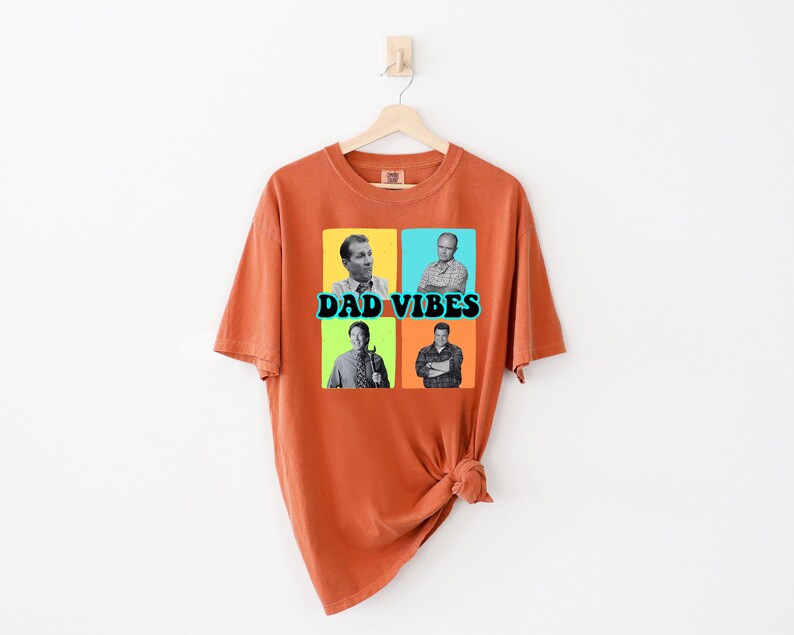 May include: Burnt orange t-shirt with a graphic that says "DAD VIBES". The graphic features four black and white images of men in a grid pattern. The shirt is hanging on a wooden hanger.