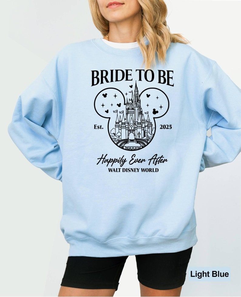 Puede incluir: Sudadera azul claro con un gr&aacute;fico negro del castillo de Cenicienta dentro de una silueta de oreja de Mickey Mouse. El texto "Bride to Be" est&aacute; por encima del gr&aacute;fico y "Happily Ever After" est&aacute; por debajo del gr&aacute;fico. El texto "Est. 2025" est&aacute; en el gr&aacute;fico.