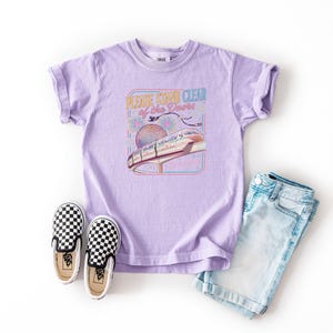 May include: A lilac t-shirt with a graphic that reads "PLEASE STAND CLEAR of the Doors" featuring a monorail and a sphere. The shirt is paired with denim shorts and black and white checkered slip-on shoes.