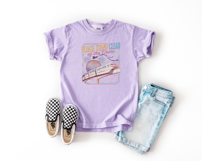May include: A lilac t-shirt with a graphic that reads "PLEASE STAND CLEAR of the Doors" featuring a monorail and a sphere. The shirt is paired with denim shorts and black and white checkered slip-on shoes.