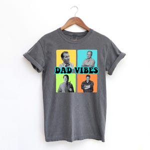 May include: Dark gray t-shirt with the words "DAD VIBES" in teal and black. The shirt features four squares with images of men in each square. The squares are yellow, blue, green, and orange.