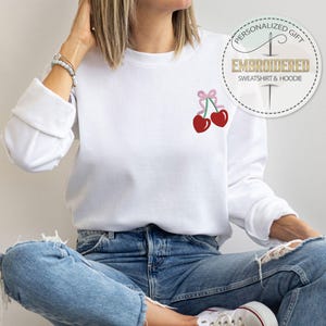 May include: White sweatshirt with a pink bow and two red cherries embroidered on the front. The text "Embroidered Sweatshirt & Hoodie" is on a circle with a needle and thread design.