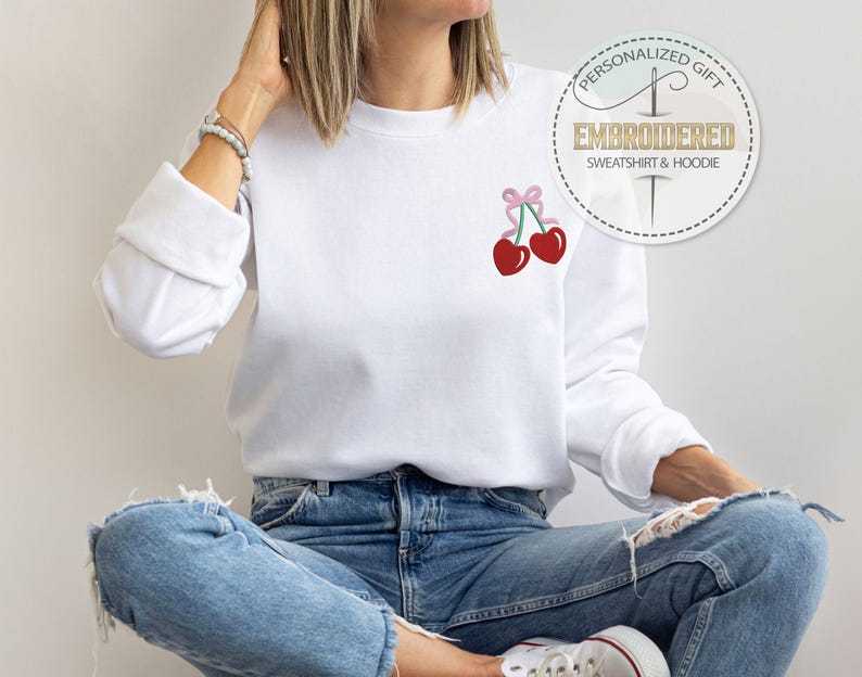 May include: White sweatshirt with a pink bow and two red cherries embroidered on the front. The text "Embroidered Sweatshirt & Hoodie" is on a circle with a needle and thread design.