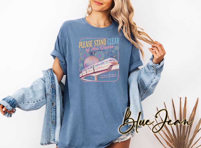 May include: Blue t-shirt with a graphic featuring a monorail, the text "PLEASE STAND CLEAR of the DOORS", and a stylized depiction of a globe. The shirt is a light blue color.