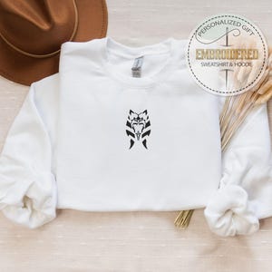 May include: White sweatshirt with a black embroidered design of a stylized face with horns. The text "Embroidered Sweatshirt & Hoodie" is on a gold circle with a needle and thread design.