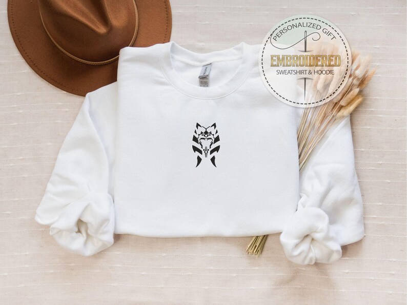May include: White sweatshirt with a black embroidered design of a stylized face with horns. The text "Embroidered Sweatshirt & Hoodie" is on a gold circle with a needle and thread design.