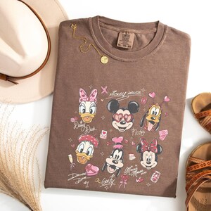 May include: A brown t-shirt featuring Disney characters Mickey Mouse, Minnie Mouse, Donald Duck, Daisy Duck, Goofy, and Pluto. The shirt is paired with a beige hat, gold necklace, and brown sandals. The shirt has the text "Mickey Mouse".