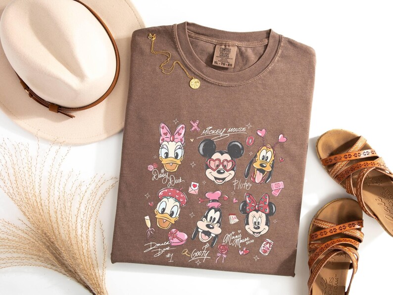 May include: A brown t-shirt featuring Disney characters Mickey Mouse, Minnie Mouse, Donald Duck, Daisy Duck, Goofy, and Pluto. The shirt is paired with a beige hat, gold necklace, and brown sandals. The shirt has the text "Mickey Mouse".