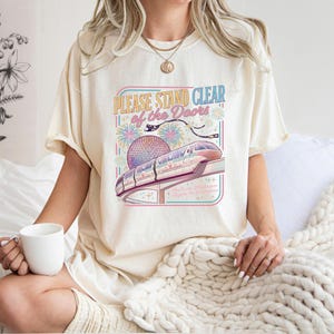 May include: Cream-colored t-shirt with the text "PLEASE STAND CLEAR of the DOORS" and a colorful graphic of a monorail and fireworks. The shirt is a relaxed fit and has short sleeves. A white mug is held in the hand.