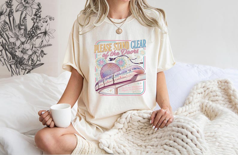 May include: Cream-colored t-shirt with the text "PLEASE STAND CLEAR of the DOORS" and a colorful graphic of a monorail and fireworks. The shirt is a relaxed fit and has short sleeves. A white mug is held in the hand.