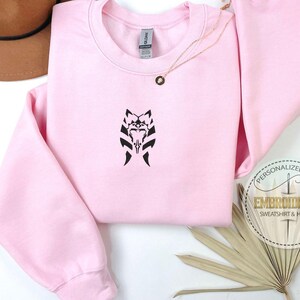 May include: Pink sweatshirt with a black embroidered design of a Mandalorian symbol. The sweatshirt is a personalized gift.