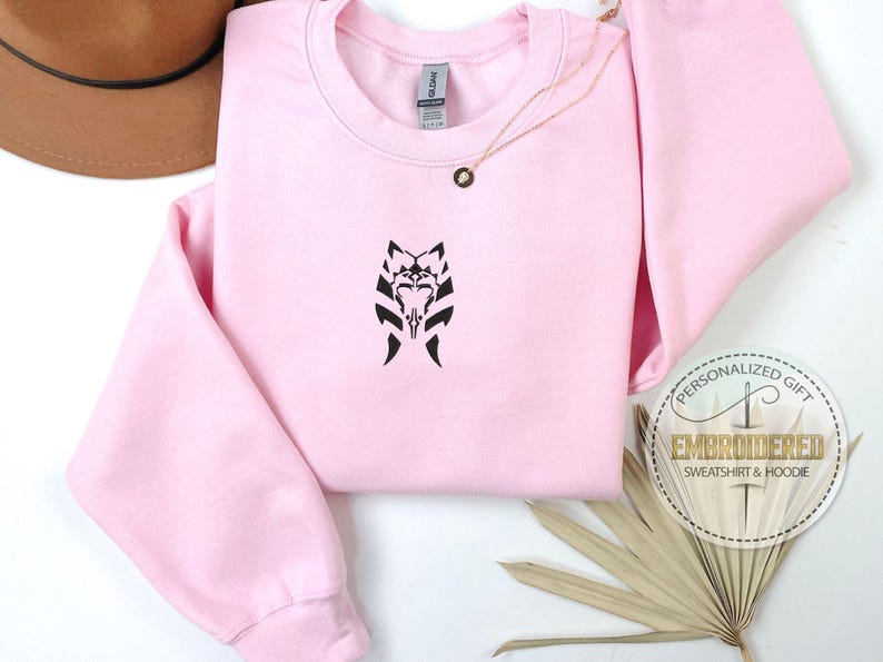 May include: Pink sweatshirt with a black embroidered design of a Mandalorian symbol. The sweatshirt is a personalized gift.