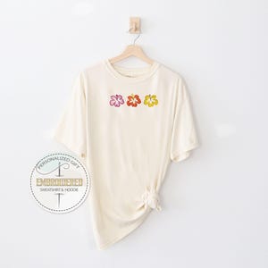 May include: Cream-colored t-shirt with three embroidered hibiscus flowers in pink, red, and yellow. The shirt is hanging on a wooden hanger against a white background. The shirt has a knotted detail at the bottom.