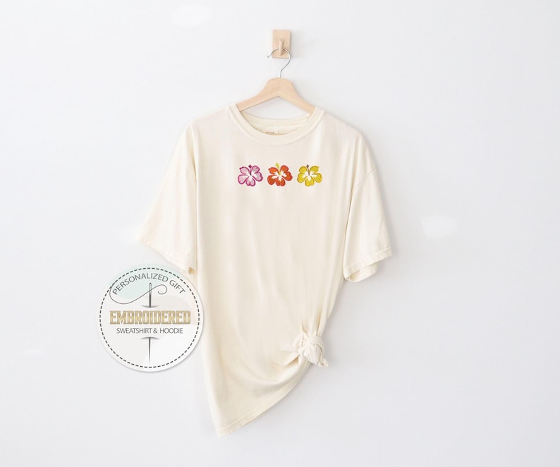 May include: Cream-colored t-shirt with three embroidered hibiscus flowers in pink, red, and yellow. The shirt is hanging on a wooden hanger against a white background. The shirt has a knotted detail at the bottom.