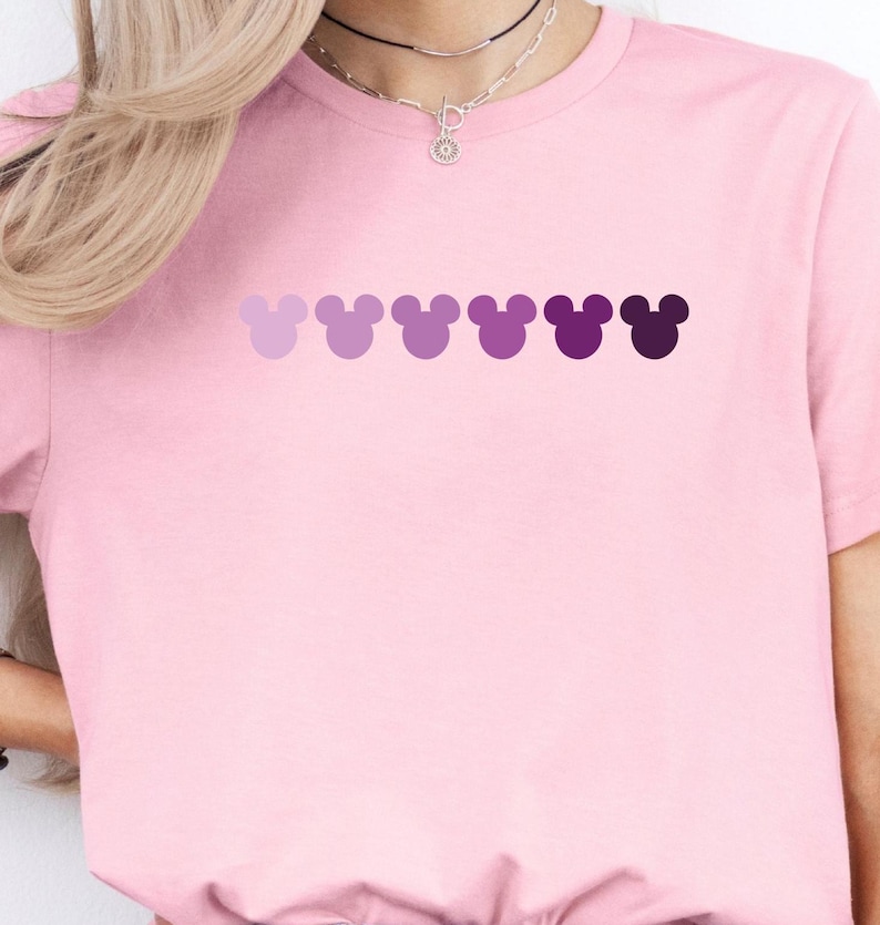Puede incluir: Una camiseta rosa con una fila de cabezas de Mickey Mouse moradas en degradado.