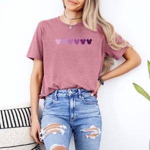Puede incluir: Una camiseta color malva con una fila de cinco cabezas de Mickey Mouse moradas en un patr&oacute;n degradado.
