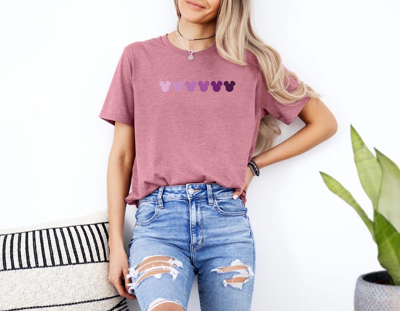 Puede incluir: Una camiseta color malva con una fila de cinco cabezas de Mickey Mouse moradas en un patr&oacute;n degradado.