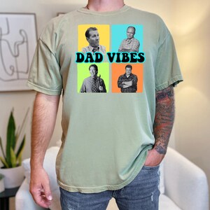 May include: Sage green t-shirt with a graphic that says "DAD VIBES" in black text. The graphic features four squares with images of men in each square. The shirt is made of a soft material.