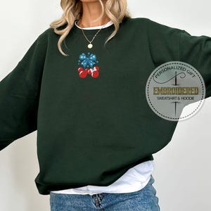 Op de afbeelding: Donkergroene sweatshirt met een witte bies en een klein geborduurd ontwerp van een blauwe strik en twee rode ornamenten. Het sweatshirt heeft de tekst "PERSONALIZED GIFT EMBROIDERED SWEATSHIRT & HOODIE" in een cirkelvormig ontwerp.