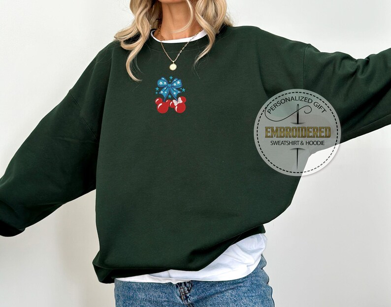 Op de afbeelding: Donkergroene sweatshirt met een witte bies en een klein geborduurd ontwerp van een blauwe strik en twee rode ornamenten. Het sweatshirt heeft de tekst "PERSONALIZED GIFT EMBROIDERED SWEATSHIRT & HOODIE" in een cirkelvormig ontwerp.