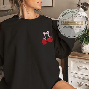 May include: Black sweatshirt with a pink ribbon and two red cherries embroidered on the left chest. The sweatshirt is a personalized gift.