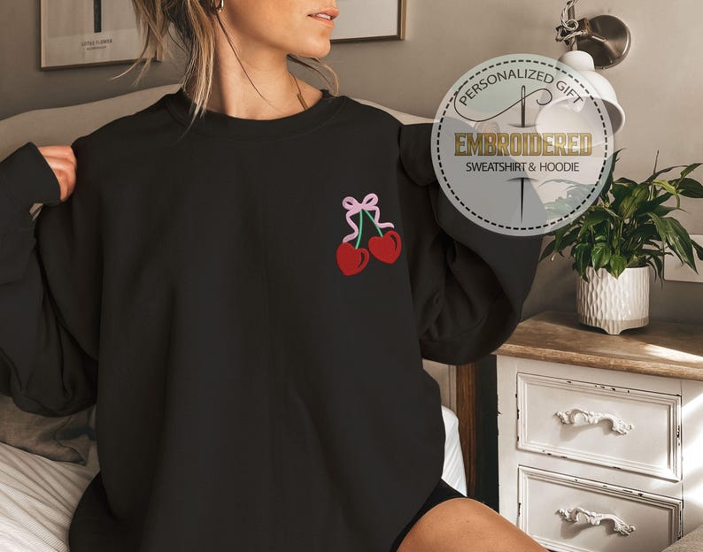 May include: Black sweatshirt with a pink ribbon and two red cherries embroidered on the left chest. The sweatshirt is a personalized gift.