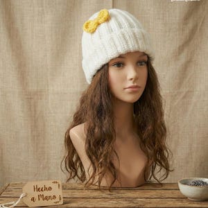May include: A white knit beanie with a yellow bow detail. The hat is displayed on a mannequin head with long brown hair. The background is a neutral fabric. A wooden tag with the text "Hecho a Mano" is visible.