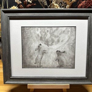 May include: A framed black and white pencil drawing of two rabbits looking up at a full moon. The drawing is on a white background and is framed in a dark gray frame.