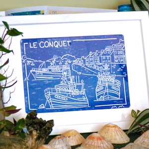 Puede incluir: Una impresión enmarcada en azul y blanco con el texto "LE CONQUET" y representaciones de barcos en un puerto. La obra de arte está en un marco blanco, rodeada de plantas y conchas marinas. La impresión tiene un tema náutico.