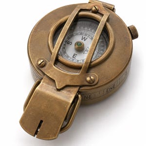 May include: A round brass compass with a weathered finish featuring a hinged lid and a protective metal frame over the glass face. The internal dial displays cardinal directions including W, S, and E, with numerical degree markings visible.