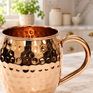 May include: A polished copper mug featuring a distinct hammered texture across its exterior surface. The mug has a wide, open top and a smooth, curved handle attached to the side, reflecting light across its metallic, warm-toned finish.