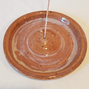 May include: A handmade ceramic incense holder with a warm, earthy color palette. The round dish features a spiral design in shades of brown and white, with a central hole for the incense stick. The holder is a functional and decorative item.