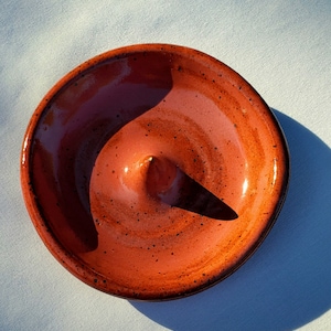 May include: A handmade, round ceramic dish with a raised center cone. The dish is glazed in a warm, reddish-brown color with darker speckles and a glossy finish. The rim is slightly darker, creating a contrast.