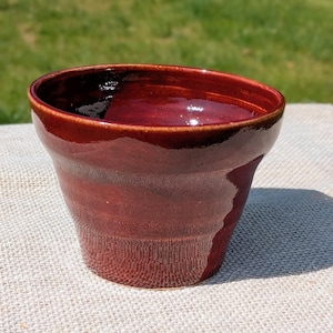 May include: A small, flared ceramic cup with a glossy, deep red glaze. The exterior surface features subtle, horizontal ridges along the lower section, while the interior is smooth and dark, reflecting light on the rim and inner walls.