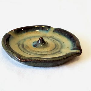 May include: A small, handmade ceramic incense holder. The dish is round with a raised center and a dark brown rim. The interior has a swirl pattern in shades of tan, blue, and green. The incense holder is designed to hold incense sticks.