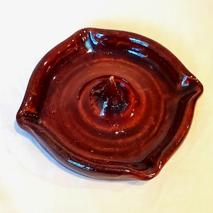 May include: A glazed, dark brown ceramic ashtray with a central cone and a circular design. The ashtray has a glossy finish and a slightly irregular, rounded shape. The interior features concentric circles, adding depth to the design.