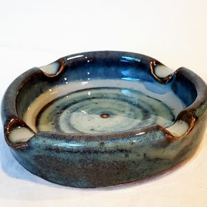 May include: A handmade ceramic ashtray with a round shape and a deep bowl. The ashtray features a blend of blue and teal glazes with brown accents. It has three curved rests for cigarettes.