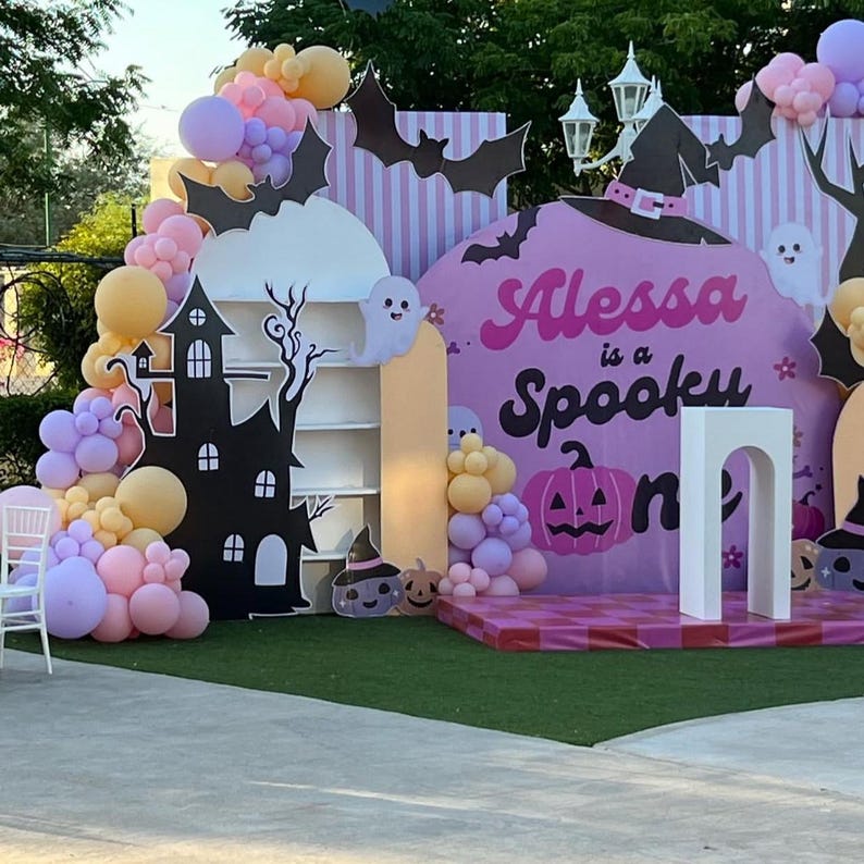 May include: A Halloween-themed party backdrop with a pink and purple color scheme. The backdrop features a black haunted house, bats, ghosts, and the text "Alessa is a Spooky One". Balloons in shades of pink, purple, and yellow decorate the scene.