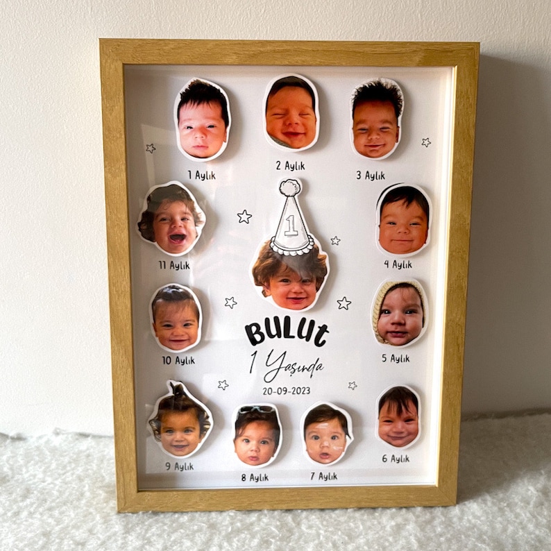 May include: Wooden frame displays a child's monthly photos from 1 month to 11 months old. Each photo is a close-up of the baby's face, and each photo is labeled with the age in months. The center of the frame shows a photo of the child at 1 year old, wearing a party hat. The text on the frame says 'BULUT 1 Yaşında 20-09-2023' which translates to 'BULUT 1 Year Old 20-09-2023'.