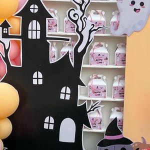 May include: A black haunted house cutout with white window details and a decorative tree. A shelf displays pink gift boxes with the text "Spooky One". A smiling ghost and a purple pumpkin with a witch hat are also present, creating a Halloween theme.